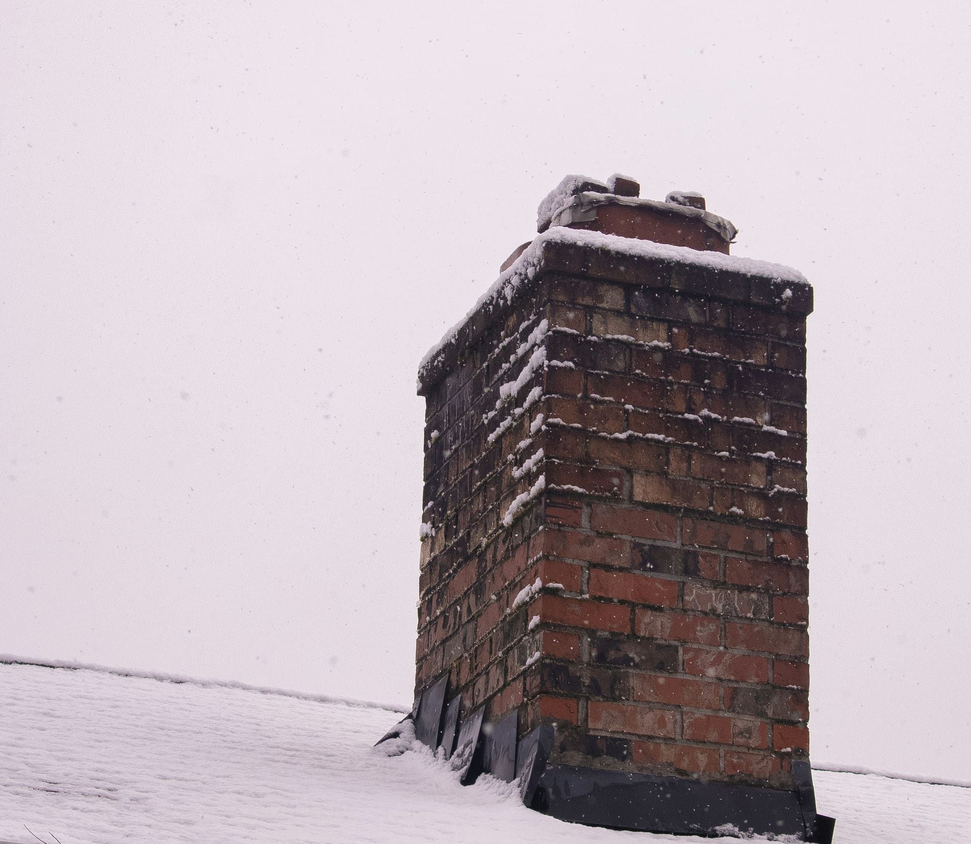 Red brick chimney on a snow-covered roof, with snow packed on its crown and drifted against the flashing at its base