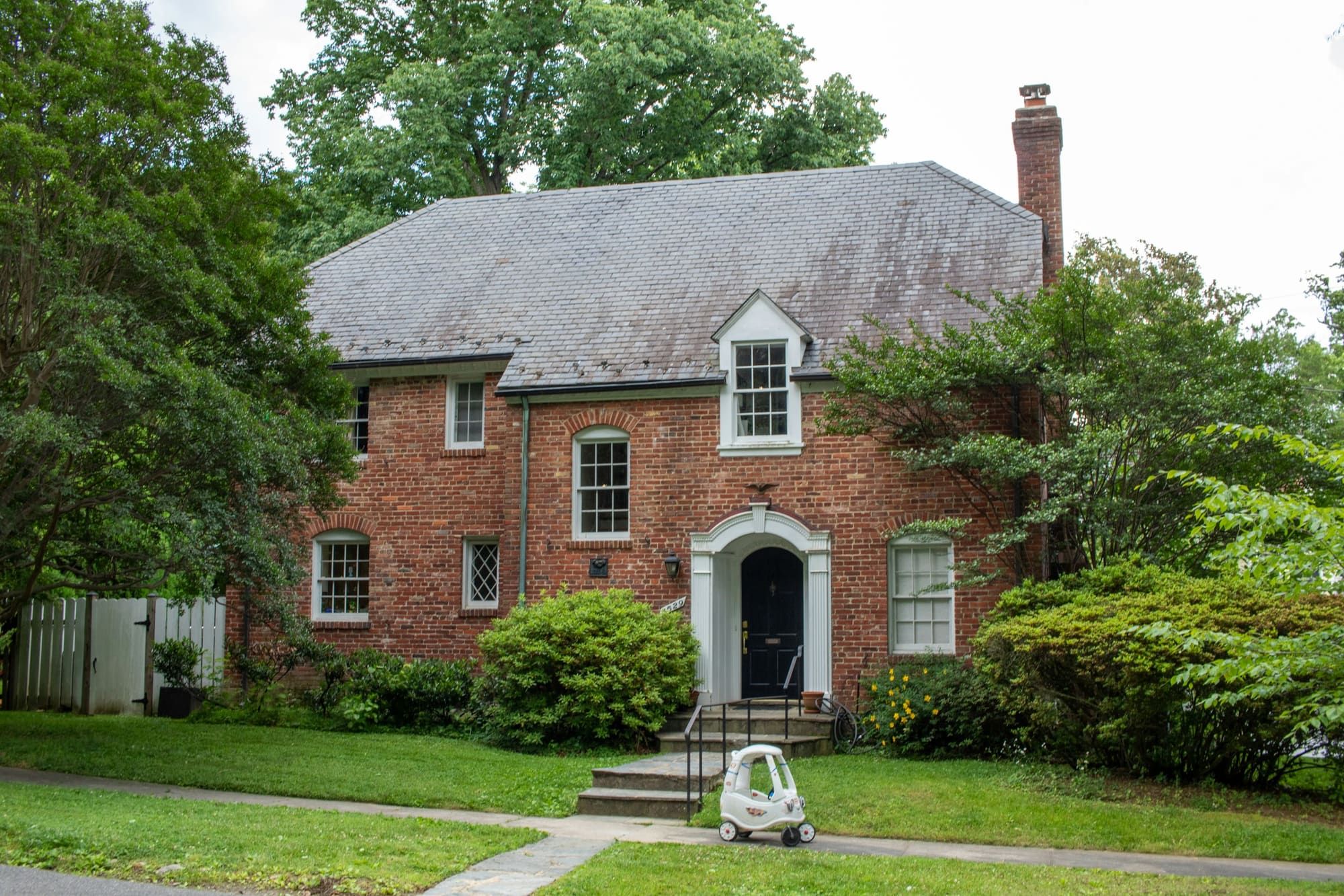 Two-story red brick house with a slate roof, a dormer and a tall brick chimney at the gable end, set behind a green lawn and mature trees