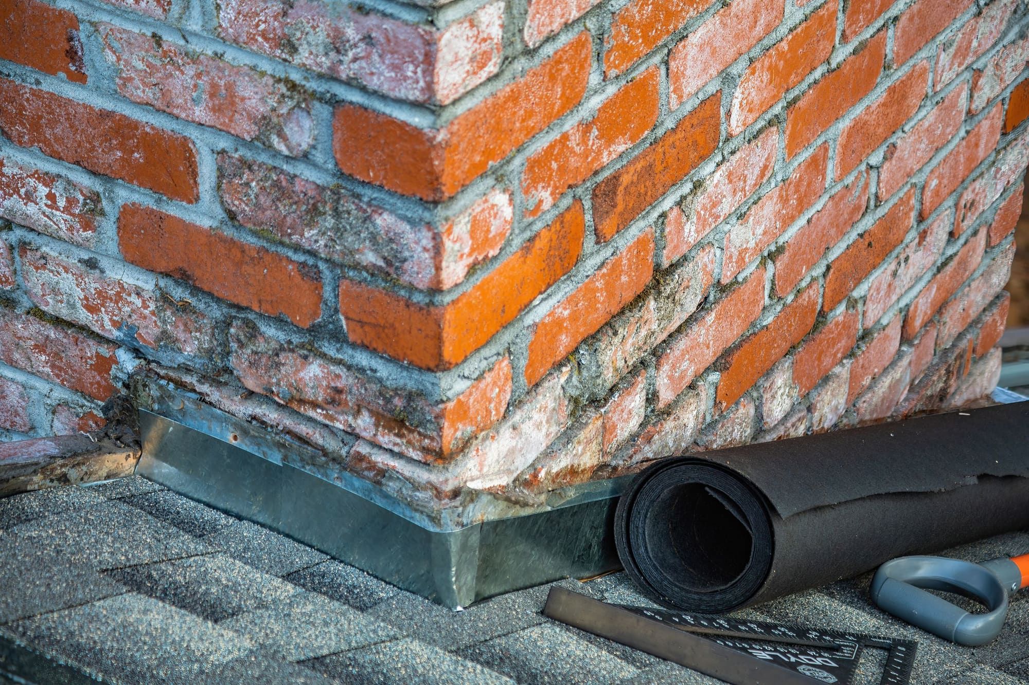 Corner of a weathered red brick chimney where it meets an asphalt shingle roof, with metal base flashing and a roll of roofing underlayment beside it