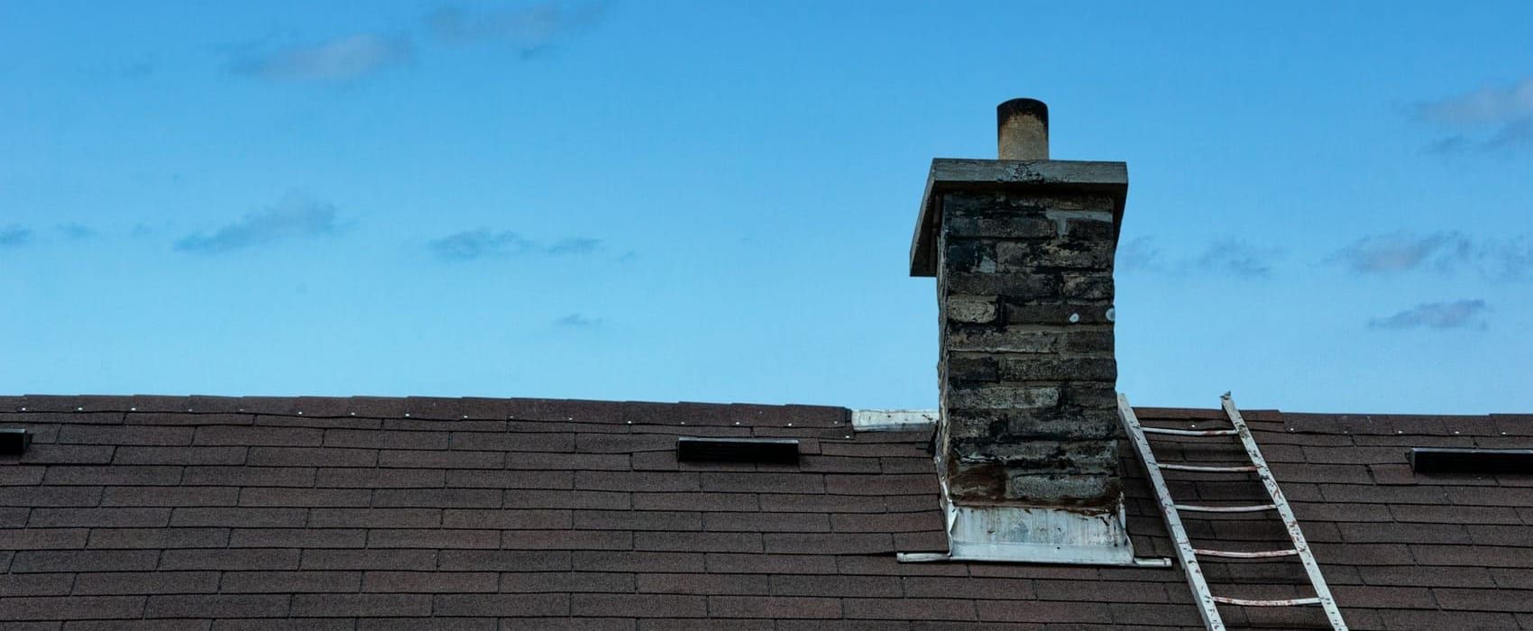 Roof ladder laid up a brown shingle roof to a weathered masonry chimney with a concrete crown and metal flashing at its base, under a clear blue sky