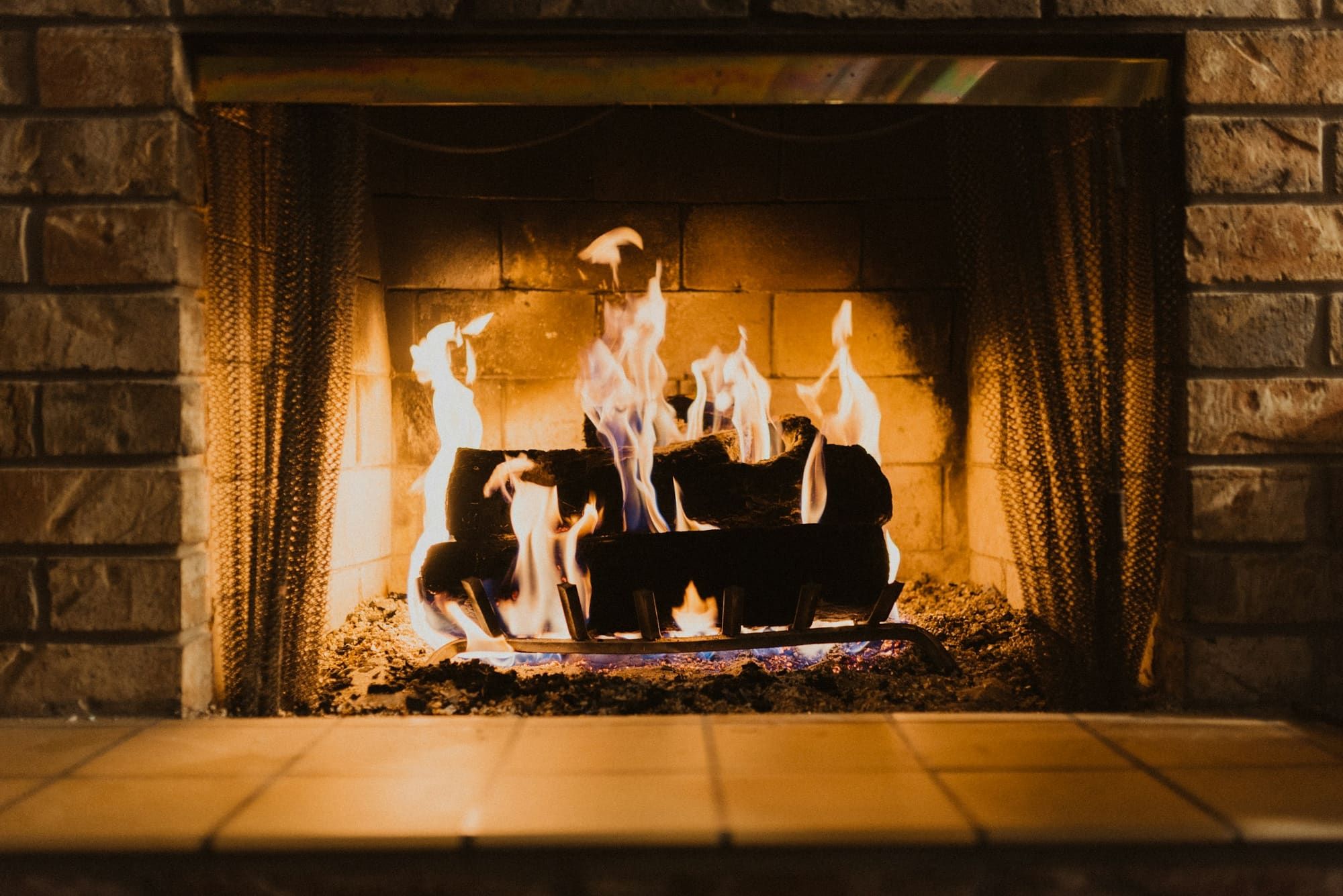 Wood fire burning on a grate inside a masonry fireplace, lighting the firebrick back wall, with a mesh spark curtain drawn open and a masonry surround on both sides