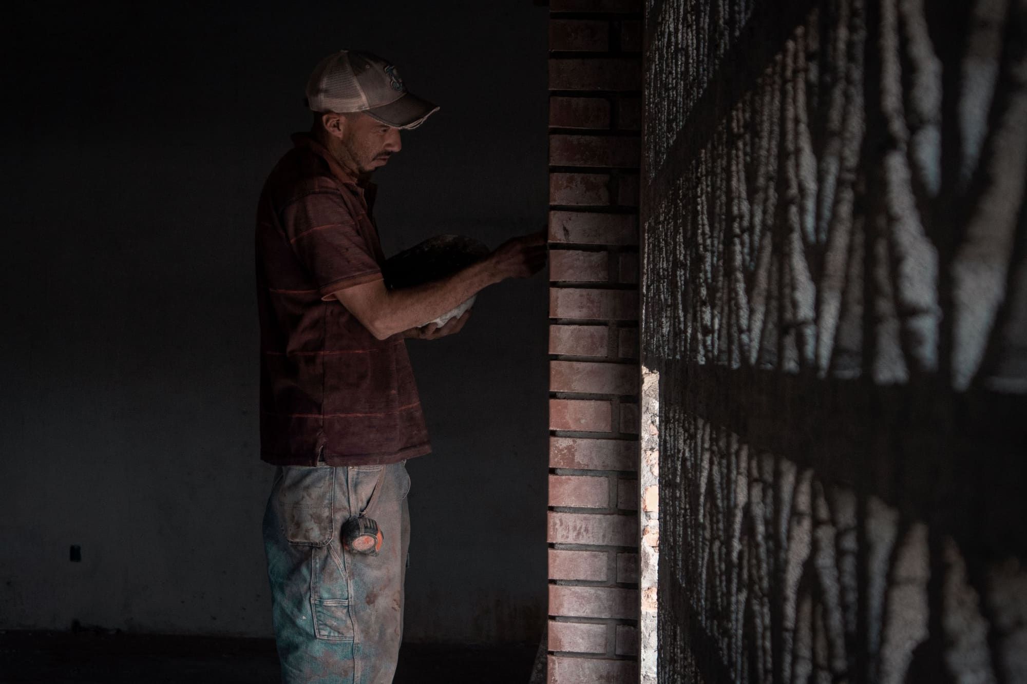 Mason in a dusty work shirt and cap holding a pan of mortar while working the joints of a red brick pier