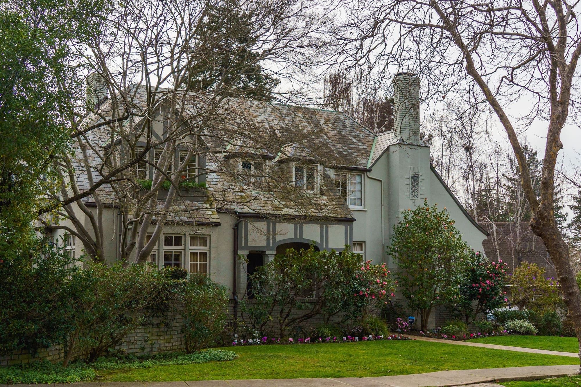 Older two-story house with gray-green stucco walls, a stucco chimney, half-timbered trim over the entry and a slate roof, seen through bare tree branches