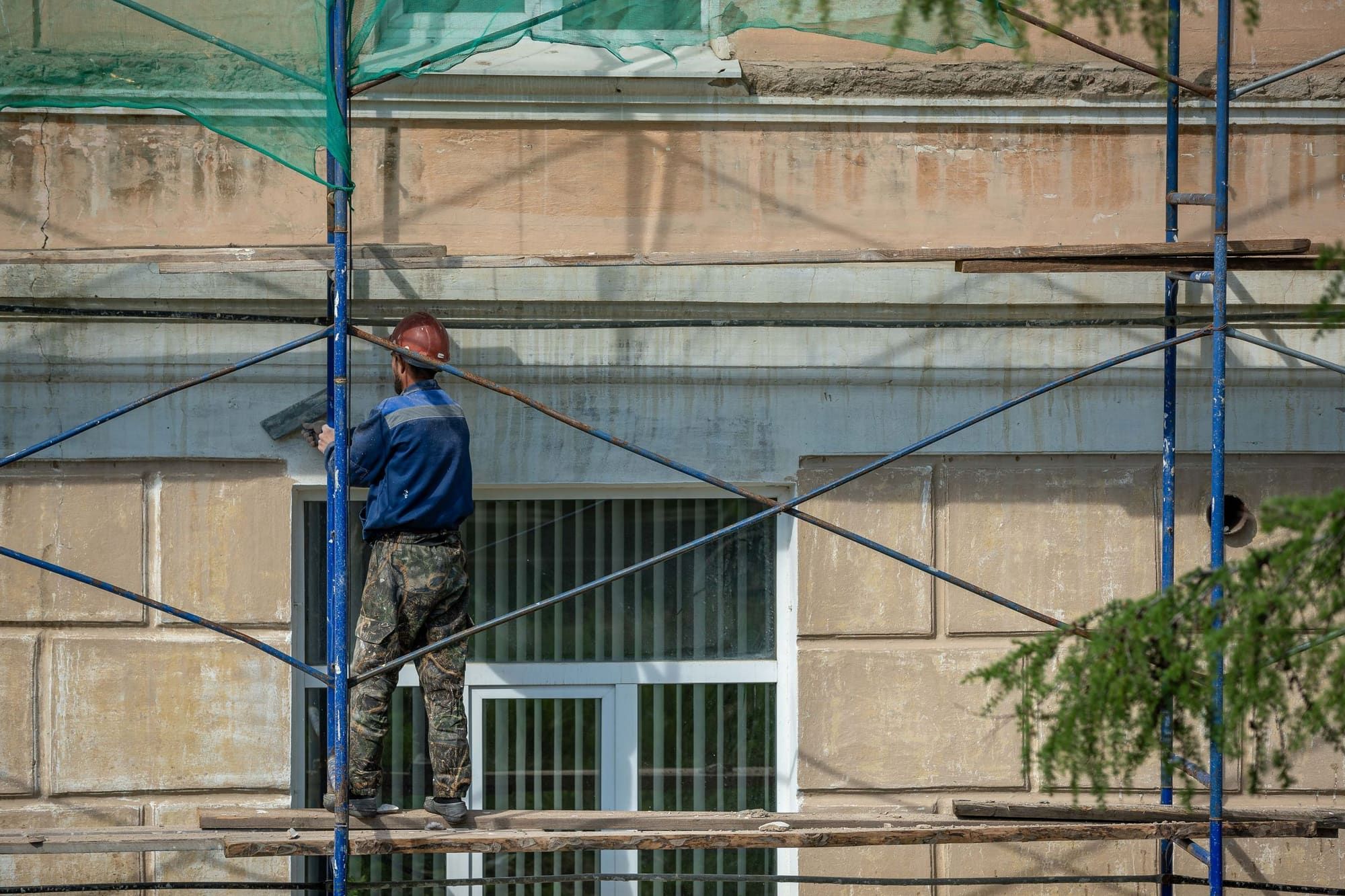 Worker in a hard hat standing on scaffold planks, seen from behind, working a float across the weathered stucco band above a window